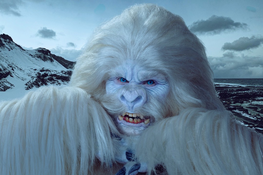 Close-up of a person in a white furry costume with a humanoid face, blue eyes, and bared teeth, set against a snowy mountain backdrop.